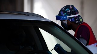 A health worker wearing a face shield at an outdoor vaccination site holds a syringe and leans in toward a person in a stopped car.