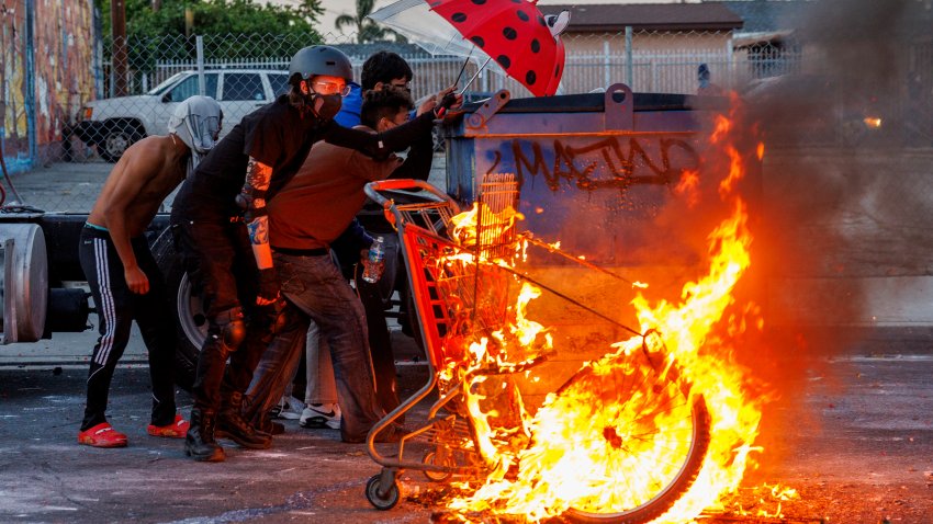 COMPTON, CA -JUNE 7, 2025: Protesters hide behind a dumpster near a set fire as LA County Sheriff deputies shoot projectiles to keep demonstrators from advancing after ICE raids at a nearby Home Depot and the Garment District brought out resistance from Los Angeles residents on June 7, 2025 in Compton, California.(Gina Ferazzi / Los Angeles Times via Getty Images)